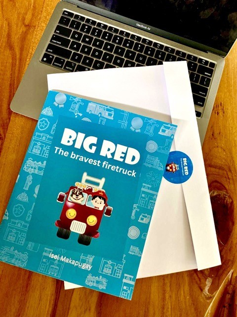 A printed copy of Big Red, The Bravest Firetruck, on a desk beside a laptop and a mailing envelope